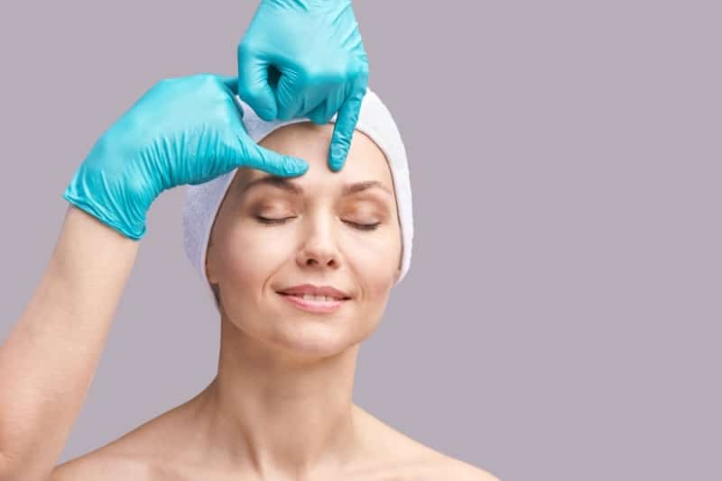 A young woman wearing a hair cap while a doctor in latex gloves examines her forehead.